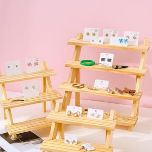 May include: Wooden tiered display stands showcasing various earrings on white cards. The earrings include studs, hoops, and drop styles, with some featuring bows and other decorative elements. A green bracelet and other jewelry are also displayed.