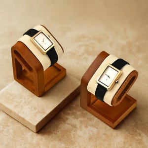 Puede incluir: Dos relojes exhibidos en soportes de madera. Cada reloj tiene una caja rectangular dorada, una esfera blanca y una correa negra. Los relojes se presentan sobre un cojín color crema dentro de un soporte de madera.