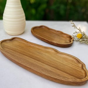 Può includere: Due vassoi di legno da portata con finitura naturale venata. I vassoi hanno un design unico con bordi ondulati. Un vaso color crema è sullo sfondo, insieme a un piccolo bouquet di fiori secchi.