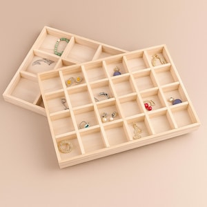 Könnte beinhalten: Zwei hellfarbene Schmucktabletts aus Holz mit mehreren Fächern. Ein Tablett ist teilweise mit verschiedenen Ohrringen, Ringen und Armbändern gefüllt. Das andere Tablett ist leer. Die Tabletts sind auf einer hellfarbigen Oberfläche angeordnet.
