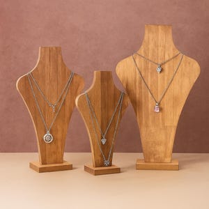 Puede incluir: Tres expositores de joyería de madera que muestran collares de plata. Cada expositor contiene varios collares con diferentes diseños de colgantes, incluyendo una flor, un corazón y una piedra preciosa. Los expositores están sobre un fondo neutro.