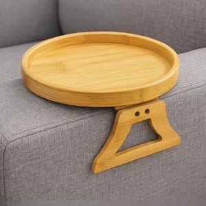 May include: A light-coloured bamboo sofa arm tray. The tray has a circular surface and a supporting bracket that fits over the arm of a grey sofa. The tray is designed to hold drinks, snacks, or other items.