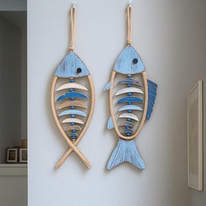 Könnte beinhalten: Zwei Wandbehänge in Form von Holzfischen. Jeder Fisch ist hellblau mit einem Naturholzrahmen und dekorativen Elementen. Die Fische sind an einem Naturseil aufgehängt. Die Wand ist hellgrau gestrichen.