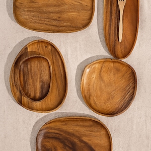 May include: A selection of wooden serving platters and bowls in different shapes. The platters are rectangular, oval, and round, with a natural wood grain finish. A wooden fork rests in one of the oval platters. Suitable for serving appetizers or small dishes.