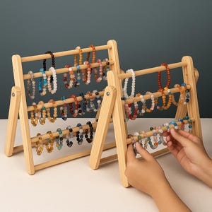 May include: Two wooden abacus-style bracelet displays. Each display has three horizontal rods holding various beaded bracelets in shades of blue, brown, orange, and white. The displays are made of light-colored wood and are set against a neutral background.