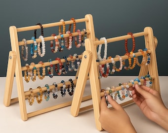 Könnte beinhalten: Zwei Armband-Displays im Abakus-Stil aus Holz. Jedes Display hat drei horizontale Stäbe, die verschiedene Armbänder mit Perlen in Blau-, Braun-, Orange- und Weißtönen halten. Die Displays sind aus hellem Holz gefertigt und vor einem neutralen Hintergrund platziert.