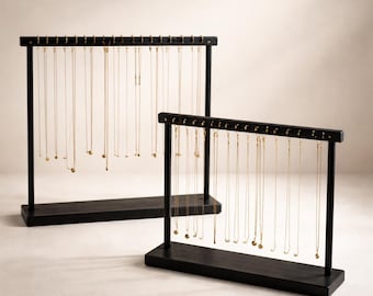 Op de afbeelding: Twee zwarte houten ketting display stands. Elke standaard heeft een rechthoekige basis en een horizontale balk met kleine pinnen om kettingen op te hangen. Gouden kettingen van verschillende lengtes worden op de standaards getoond.
