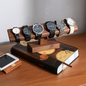 Peut inclure: Un présentoir à montres en bois avec six montres de différents styles de bracelets. Les montres ont des cadrans noirs, argentés et blancs. Le présentoir est posé sur un livre décoratif et un petit portefeuille et une tablette en cuir sont sur la table.