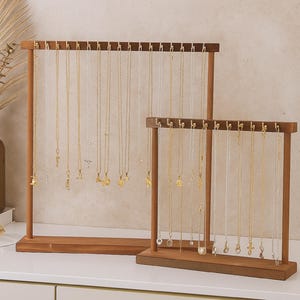 May include: Two wooden necklace display stands. The larger stand holds numerous necklaces with various pendants. The smaller stand also displays necklaces. Both stands are made of brown wood and sit on a white surface.