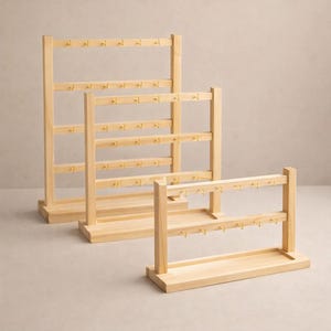 May include: Three wooden jewelry display stands of varying heights. Each stand has multiple horizontal bars with small hooks for hanging earrings. The stands are made of light-colored wood and have a simple, minimalist design.