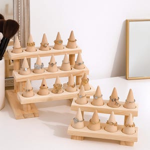 Peut inclure: Présentoirs à bagues en bois à plusieurs niveaux, présentant diverses bagues. Les présentoirs ont une finition en bois naturel et comportent des supports en forme de cône pour chaque bague. Les bagues sont de tons or et argent, avec différents modèles.
