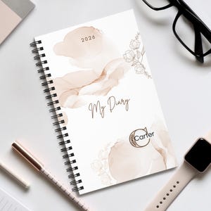 Peut inclure: Un agenda à spirales avec une couverture blanche ornée de motifs abstraits marron et beige façon aquarelle. Le texte "My Diary" est écrit en cursive, avec l'année "2026" et le nom de la marque "Carter". Un stylo, des lunettes et une montre connectée sont également visibles.