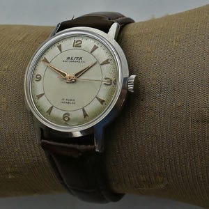 Op de afbeelding: Een vintage Blita polshorloge met een zilverkleurige ronde kast, een crèmekleurige wijzerplaat met goudkleurige wijzers en uurmarkeringen, en de tekst Blita Antimagnetic 17 Rubis Incabloc. Het horloge heeft een donkerbruine lederen band met een textuur.