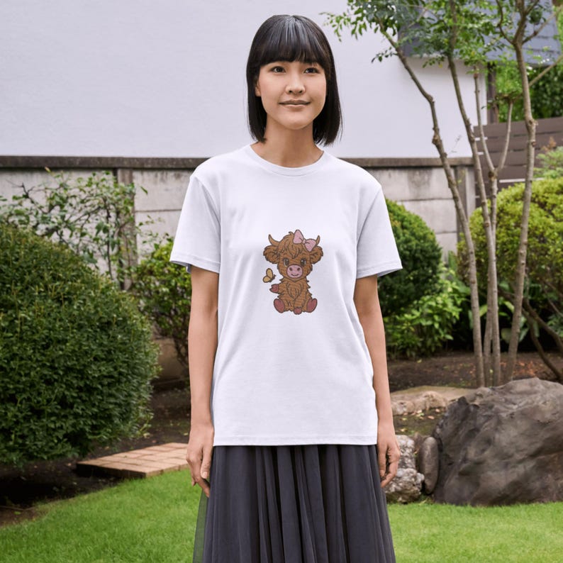 Op de afbeelding: Wit T-shirt met een illustratie van een Schotse Hooglander. De koe is bruin met een roze strik en een vlinder. Het T-shirt is gemaakt van zacht materiaal. De koe zit. Het T-shirt is wit.
