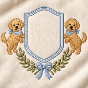 May include: An off-white fabric featuring an embroidered design of two golden-coloured puppies wearing light blue bandanas. The puppies flank a central light blue shield-shaped frame, positioned above a green laurel wreath and a light blue bow.