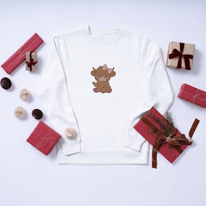 Op de afbeelding: Wit sweatshirt met een geborduurde cartoon Highland koe met een roze strik. De koe is bruin met een lichtroze strik. Verschillende verpakte cadeaus in rood en goud zijn verspreid.