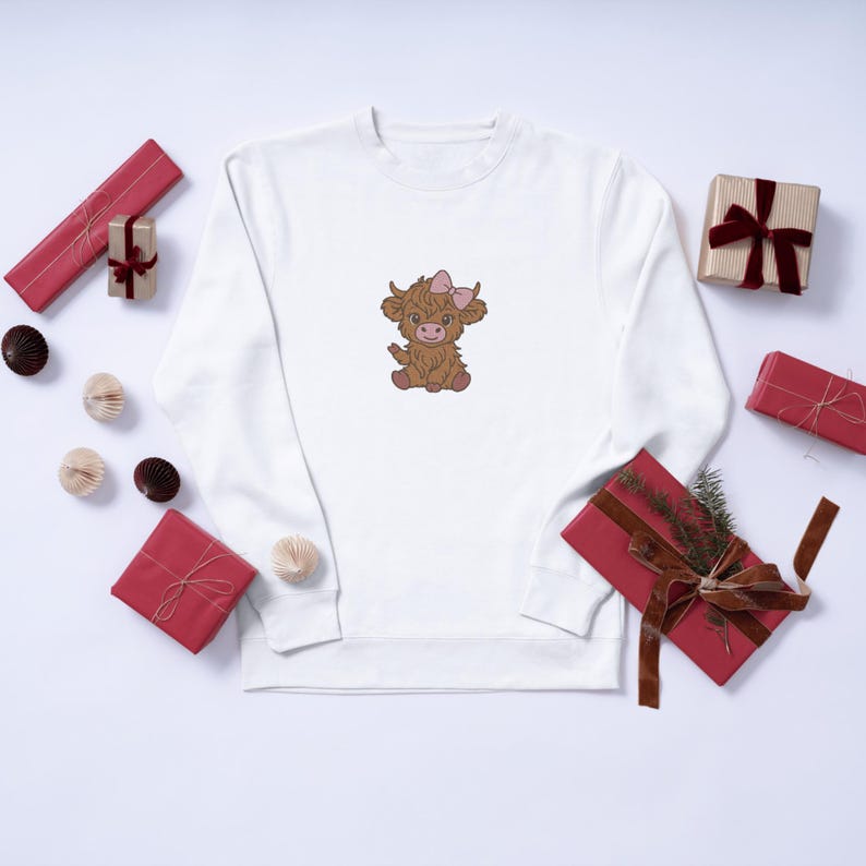 Op de afbeelding: Wit sweatshirt met een geborduurde cartoon Highland koe met een roze strik. De koe is bruin met een lichtroze strik. Verschillende verpakte cadeaus in rood en goud zijn verspreid.