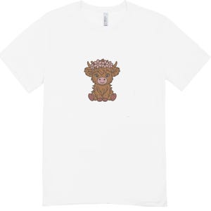 Op de afbeelding: Wit T-shirt met een cartoon illustratie van een Schotse Hooglander. De koe is bruin en draagt een roze bloemenkrans. Het ontwerp is gecentreerd. Het T-shirt is gemaakt van een zacht, comfortabel materiaal.