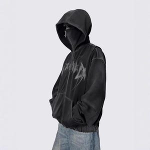 May include: Black zip-up hoodie with a hood and a face covering. The hoodie features a distressed, abstract design across the chest. The model is wearing blue jeans. The hoodie has two front pockets.