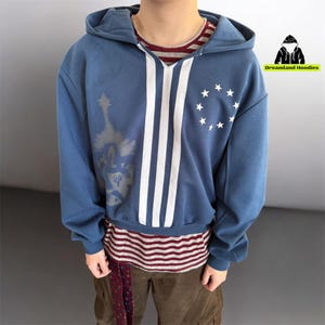 May include: Blue hooded sweatshirt with white stripes and a circle of white stars. The sweatshirt has a graphic on the left side and is layered over a striped shirt. The brand logo "Dreamland Hoodies" is visible in the upper right corner.