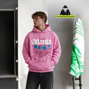 May include: Pink hoodie with the word "Atlanta" in white script, blue stars, and a spiderweb design. The hoodie has a hood and a front pocket. The brand "Dreamland Hoodies" is visible in the top right corner.