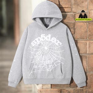 May include: A gray hooded sweatshirt with a white spider web design and the word "sp5der" across the chest. The sweatshirt has a front pocket and stars on the sleeve. The brand logo "Dreamland Hoodies" is in the upper right corner.