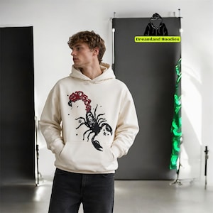 May include: Cream-coloured hoodie with a black scorpion graphic and red "BOSS" text. The hoodie has a kangaroo pocket and a drawstring hood. The brand name "Dreamland Hoodies" is visible in the background.