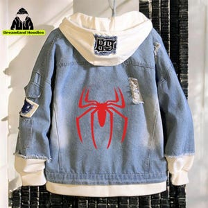 Op de afbeelding: Een lichtblauwe spijkerjas met een witte capuchonvoering en manchetten. De achterkant van de jas heeft een grote rode spinnenafbeelding. De jas heeft distressed details en patches. De tekst "Dreamland Hoodies" is zichtbaar.
