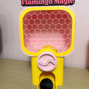 Puede incluir: Un juguete amarillo y rosa con el texto "Flamingo Magic" en la parte superior. El juguete tiene un interior con patrón de panal, un diseño circular rosa y una bola negra en la parte inferior. El juguete está diseñado para dispensar la bola negra.