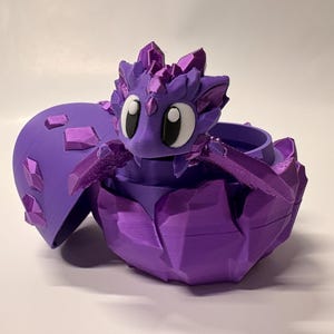Puede incluir: Un recipiente morado con forma de dragón con tapa con bisagras. El dragón tiene ojos grandes y detalles de cristal. El recipiente está abierto, revelando un interior circular. El diseño está impreso en 3D.