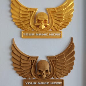 Könnte beinhalten: Zwei geflügelte Totenkopf-Embleme in Gold und Bronze. Jedes zeigt einen Totenkopf in Hufeisenform, mit dem Text "YOUR NAME HERE" auf einem rechteckigen Schild. Die Flügel sind detailliert und ausgebreitet.