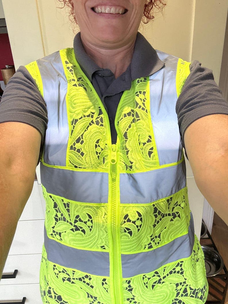 Op de afbeelding: Een veiligheidsvest met hoge zichtbaarheid met een neongeel kanten ontwerp en reflecterende zilveren strepen. Het vest heeft een lange ritssluiting en wordt over een grijs overhemd met kraag gedragen.