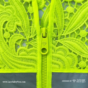 Op de afbeelding: Close-up van een neon groen veiligheidsvest met ingewikkelde kanten details. Het vest heeft een lange ritssluiting en een reflecterende grijze band aan de onderkant. De tekst "www.LaceSafetyVest.com" is zichtbaar.