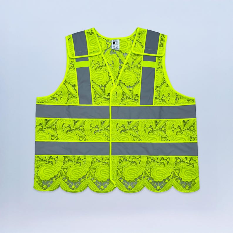 May include: A neon yellow safety vest with a floral lace pattern. The vest has gray reflective stripes and accents, with a scalloped hemline. The vest has a button closure and shoulder straps.
