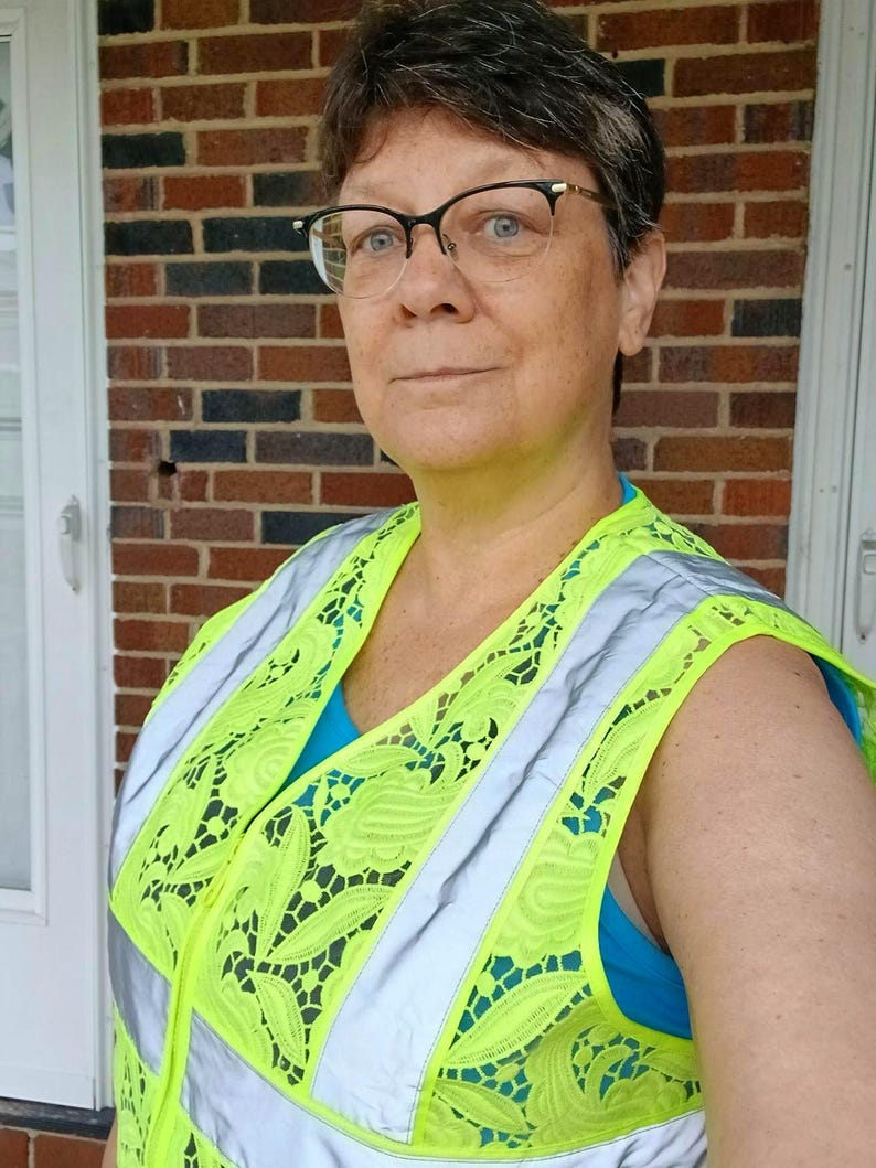 Op de afbeelding: Een felgele veiligheidsvest met reflecterende zilveren strepen en een decoratief kanten patroon. Het vest wordt gedragen over een blauwe top. De persoon draagt een bril.