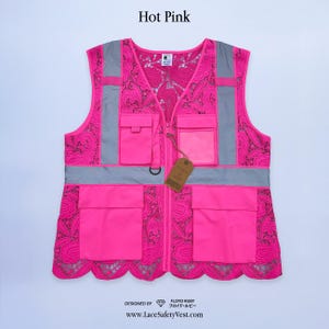 Może przedstawiać: Kamizelka odblaskowa z różowej koronki z odblaskowymi szarymi paskami i kieszeniami. Kamizelka ma falisty dół i zapięcie na zamek błyskawiczny. Tekst "Hot Pink" znajduje się na górze. Strona internetowa www.LaceSafetyVest.com znajduje się na dole.