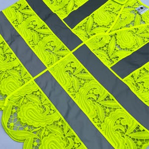 May include: A high-visibility safety vest with a unique design. The vest features bright neon yellow lace detailing with a floral pattern, contrasted by gray reflective stripes. The intricate lace adds a decorative element to the functional safety gear.