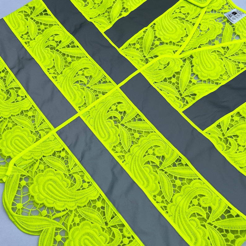 May include: A high-visibility safety vest with a unique design. The vest features bright neon yellow lace detailing with a floral pattern, contrasted by gray reflective stripes. The intricate lace adds a decorative element to the functional safety gear.