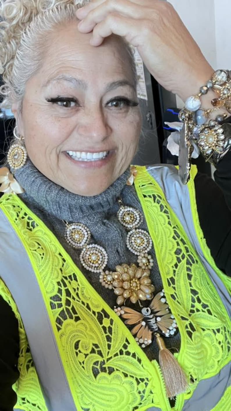 Op de afbeelding: Een vrouw draagt een grijze coltrui, een neongeel vest en uitgebreide sieraden. De sieraden omvatten een grote ketting met bloemen- en vlinderhangers, oorbellen en meerdere armbanden. Het vest heeft een kantachtig ontwerp.
