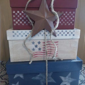 May include: A stack of three rectangular wooden boxes in red, white, and blue. The top red box has a checkered pattern, the middle white box features a stenciled American flag, and the bottom blue box has white stars. A metal star is tied on with twine.