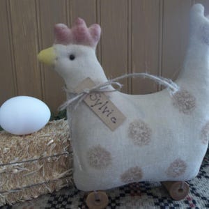 May include: A stuffed chicken toy with a pink comb, yellow beak, and brown polka dots. The chicken has a tag that says "Sylvia". A white egg rests on a small hay bale next to the chicken.