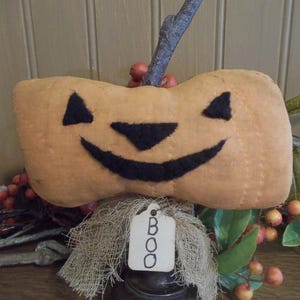 May include: A stuffed fabric pumpkin in a muted orange tone with a black felt jack-o'-lantern face. It features a natural twig stem and a burlap collar adorned with a small white paper tag that has the word BOO written in black ink.