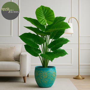 May include: A large, artificial green plant with broad leaves in a turquoise pot with a gold floral design. The pot sits on a light-colored rug. A gold floor lamp with a white shade is to the right. The logo for Silken Stems is in the upper left.