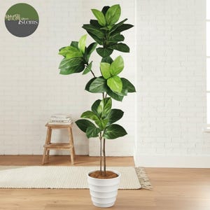 May include: A tall artificial fiddle leaf fig tree with large, vibrant green leaves. The tree is in a white, tiered pot and stands on a wooden floor. The background is a white brick wall. The logo for "Silken Stems" is in the upper left corner.