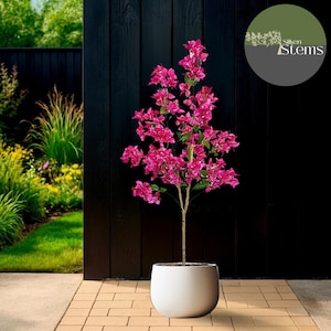 May include: A bright pink bougainvillea plant in a white, rounded pot. The artificial plant has a slender, light brown trunk and is set against a black wooden fence. The image includes the text "Silken Stems" in a circular logo.
