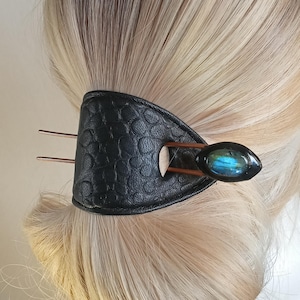 Puede incluir: Accesorio para el cabello de cuero negro con un patrón texturizado, alfileres de color cobre y una piedra preciosa ovalada azul y verde. El accesorio para el cabello está diseñado para sujetar el cabello en su lugar.