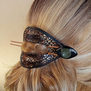 Op de afbeelding: Decoratieve haaraccessoire met een zwarte leren band, met een patroon van goud en zilverkleurige spikkels. Het heeft koperkleurige pinnen en een zwarte, ovale steen. Het haarstuk wordt getoond in blond haar.