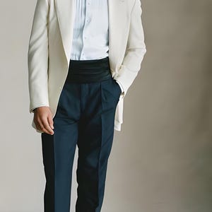 Könnte beinhalten: Ein cremefarbener Smoking mit schwarzer Fliege, hellblauem Hemd, schwarzem Kummerbund und marineblauer Anzughose. Das Outfit wird durch schwarze Anzugschuhe ergänzt, perfekt für einen besonderen Anlass.