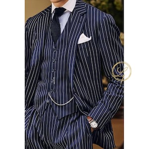 May include: Navy blue three-piece suit with white pinstripes. The suit includes a jacket, vest, and trousers. A white pocket square and a dark tie complete the look. A silver chain is attached to the vest. A wristwatch with a brown leather band is visible.