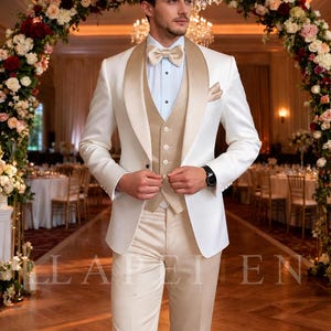 May include: A formal ivory suit with a champagne-colored lapel and vest. The suit includes a matching bow tie and pocket square. The background features floral archways and a chandelier, suggesting a wedding or formal event.
